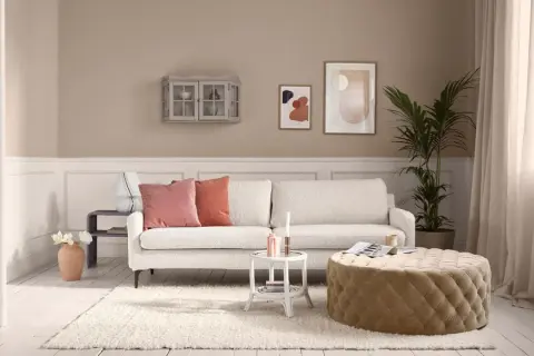 Beige ist die Farbe der Wände, des Sofas und des runden Hockers. Ein gemütliches Wohnzimmer mit weißen Dielen und einem Teppich.