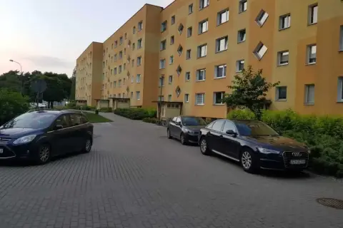 Na drodze osiedlowej zaparkowane są samochody przed blokiem z żółtą elewacją i nietypowymi oknami.