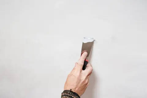 Mano con spatola applica stucco bianco su parete, un passo per come pitturare di bianco una parete gialla.