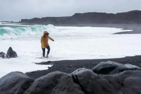 Kobieta w żółtej kurtce brodzi w pianie na czarnej plaży Reynisfjara, podczas gdy potężne fale rozbijają się o brzeg.
