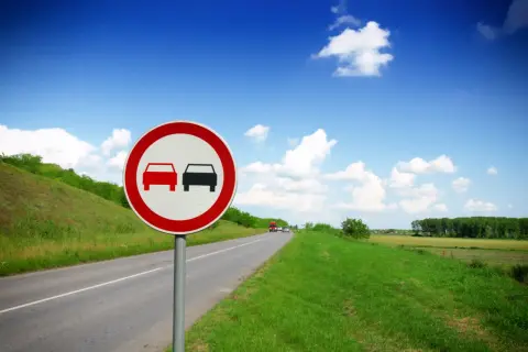 Znak zakaz wyprzedzania na malowniczej drodze wśród zielonych wzgórz i błękitnego nieba.