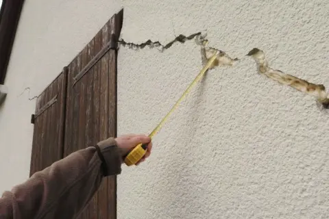 Main d'une personne mesurant une micro fissure sur un crépi extérieur. La fissure est large et profonde, révélant l'isolant.