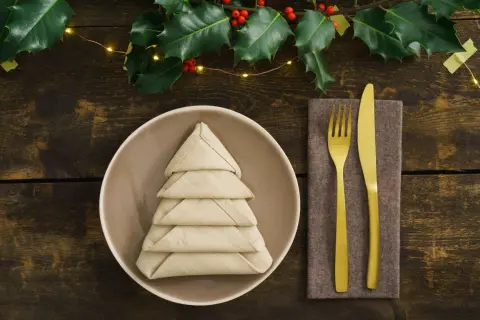 Une assiette avec un pliage serviette rectangulaire en tissu facile en forme de sapin. Couverts dorés sur une serviette marron. Branche de houx et guirlande lumineuse.
