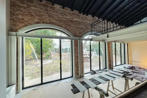 Interno in fase di ristrutturazione con ampie vetrate sul giardino. In primo piano, assi di legno appoggiate su cavalletti, segno di lavori di miglioramento della casa in corso.