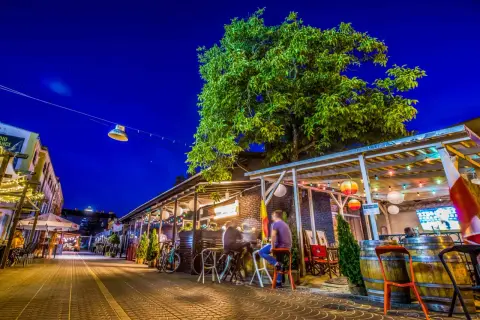 Wieczorna ulica z ogródkami restauracyjnymi, drzewem i oświetleniem.
