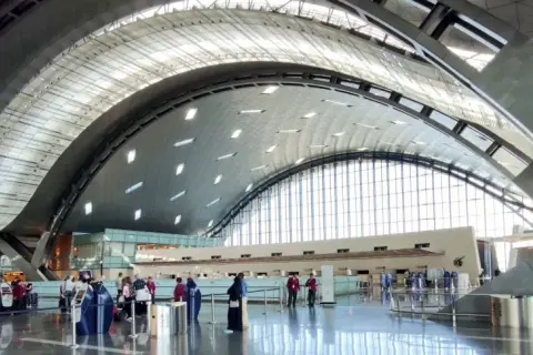 Nowoczesne wnętrze lotniska w Doha z charakterystycznym, falującym dachem i przeszklonymi ścianami. Pasażerowie czekają w kolejce do odprawy.