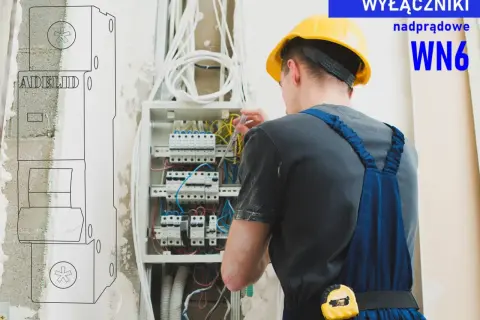 Elektryk w żółtym kasku pracuje przy rozdzielnicy, dokonując doboru zabezpieczeń nadprądowych. WN6.