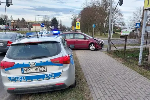 Samochód policyjny z włączonymi sygnałami świetlnymi przy drodze, gdzie doszło do kolizji drogowej. Czerwony samochód ma uszkodzony przód.