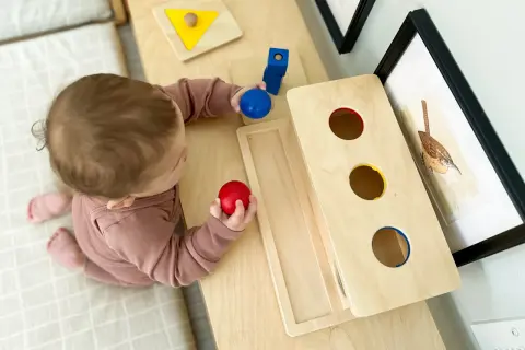 A baby explores Montessori activities for 10 month old baby, holding red and blue balls near a wooden drop box and shape puzzle.