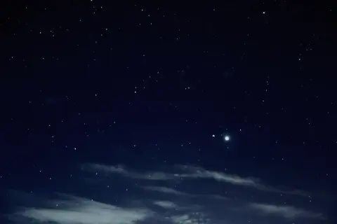 Nocne niebo pełne gwiazd, z widocznymi chmurami i dwoma jasnymi punktami, być może planetami.