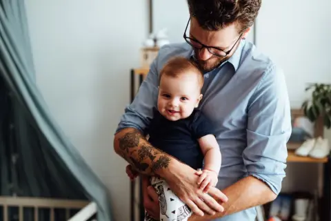 Ein Mann hält sein Baby. Auf seinem Unterarm sind Tätowierungen, die an die Namen seiner Kinder erinnern.