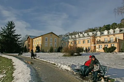 Żółty budynek sanatorium otoczony śniegiem, ludzie siedzą na ławce.