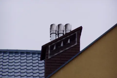 Trzy nasady kominowe na ceglanym kominie, świadczące o prawidłowym montażu nasady kominowej. Obok dach z niebieską dachówką.