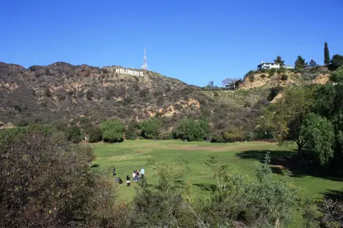 En grönskande park med människor som njuter av solen, med den ikoniska hollywood skylten i bakgrunden mot en klarblå himmel.