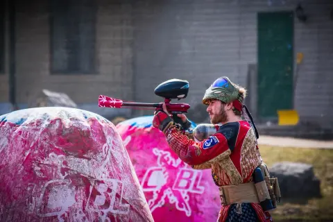 Gracz w stroju bojowym celuje z markerem, gotowy do akcji w laserowym paintballu. Za nim różowe dmuchane przeszkody.