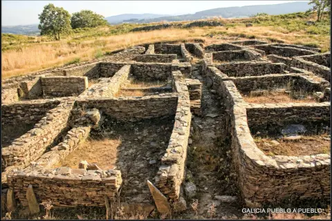 Ruinas de piedra del castro de Castromaior, con muros bajos y patios interiores, bajo un cielo azul y vegetación seca.