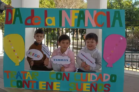 Niños sonríen tras un marco decorado para el Día de la Infancia, sosteniendo carteles con palabras como "Juego", "Familia" y "Protección".