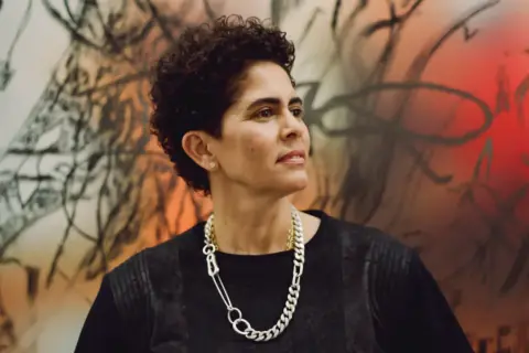 A striking portrait of artist Julie Mehretu, her gaze thoughtful, set against a vibrant, abstract background that echoes her dynamic style.