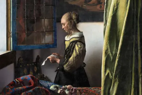 A woman in a yellow and black dress reads a letter by a window, a scene from one of Vermeer's famous paintings of women.
