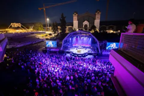 Ein belebtes **Prag Festival** mit einer großen Menge, die einer Band auf einer beleuchteten Bühne zusieht.