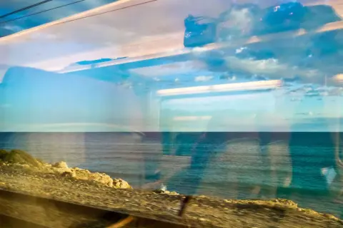 Blick aus dem Zugfenster auf das Meer. Die Spiegelungen im Glas zeigen Wolken und den Himmel, während draußen die Küste vorbeizieht. Mit dem Zug ans Meer ist ein Erlebnis.