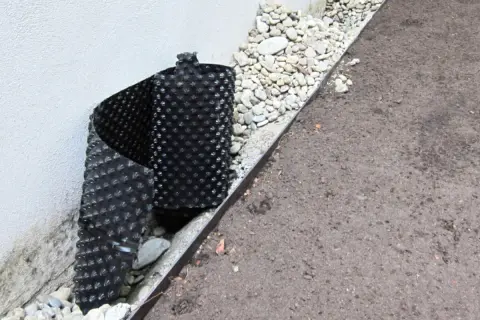 Schwarze Noppenbahn wird an einer Hauswand angebracht, um Feuchtigkeit abzuleiten. Sie klebt gut an der Wand.
