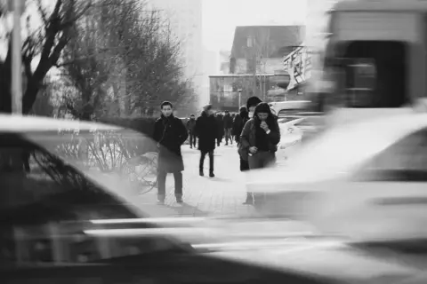 Ludzie na chodniku, rozmyci przez długi **shutter speed**. W tle drzewa i budynki.