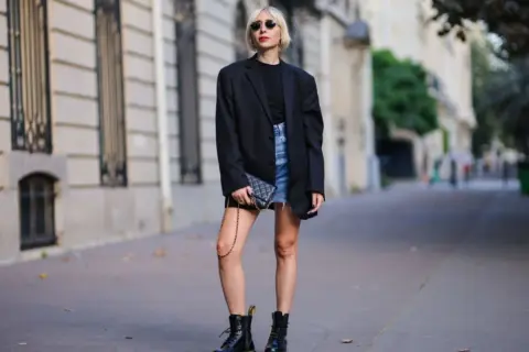 Kobieta w czarnym blazerze i jeansowej spódniczce nosi kultowe buty Dr. Martens, które mają bogatą historię.