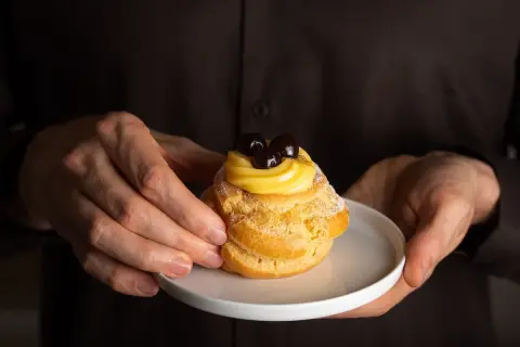 Mani che offrono una zeppola di San Giuseppe, un dolce con una ricca storia, guarnita con crema pasticcera e amarene.