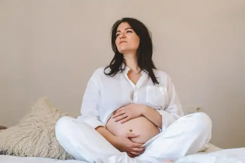 Kobieta w białej koszuli i spodniach siedzi na łóżku, obejmując brzuch. Jej zamyślona mina może sugerować tokofobia objawy.