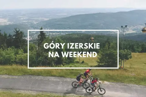 Trzech rowerzystów przemierza malownicze szlaki w Górach Izerskich w Polsce. W tle widać kolejkę gondolową i rozległy krajobraz.