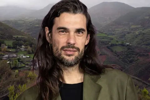 Oliver Laxe, director, posa con una barba cuidada y pelo largo, frente a un paisaje montañoso de Galicia.