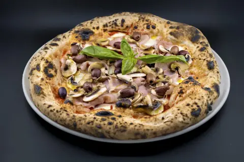Pizza Capricciosa con champiñones, jamón y aceitunas negras, coronada con albahaca fresca. ¡Un verdadero capricho!