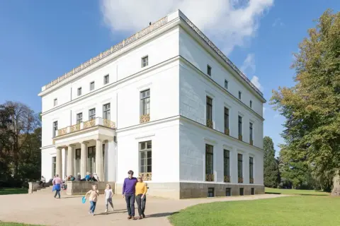 Familie spaziert durch den Jenisch Park vor einem weißen Herrenhaus mit Säulen und goldenen Verzierungen.