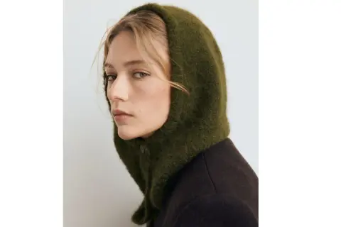 Mujer con un balaclava verde de lana, cubriendo su cabeza y cuello, con un abrigo oscuro.