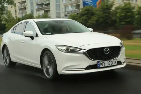 Biała Mazda 6 2.0 benzyna jedzie po mokrej drodze, w tle budynki i drzewa.