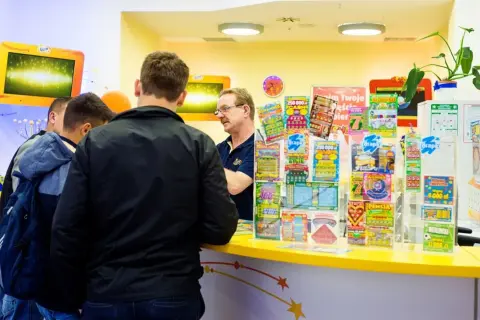 Mężczyźni stoją przy ladzie, na której widać wiele zdrapek Lotto. Czy na Orlenie są zdrapki Lotto? Tak, jak widać na zdjęciu.