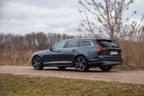 Ciemnoszary Volvo V90 T8 na drodze gruntowej, otoczony suchą trawą i drzewami.