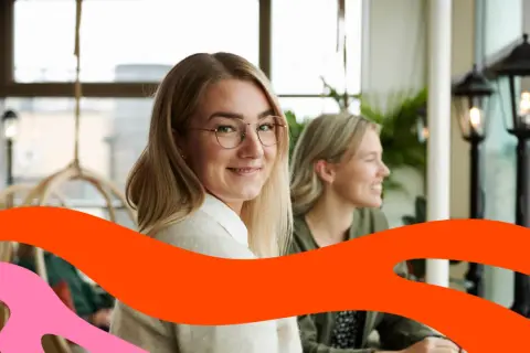 Två leende kvinnor sitter vid ett bord. Den ena tittar mot kameran. Kanske funderar de på vad som går arbetsgivaravgiften till.