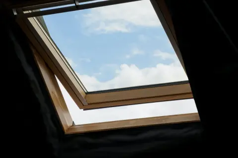 Widok na błękitne niebo z chmurami przez uchylone okno dachowe. Drewniana rama okna sugeruje możliwe rozszczelnienie.