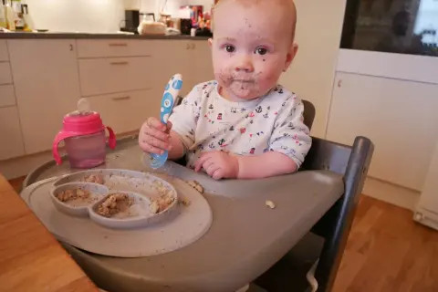 Sött litet barn med mat i ansiktet, redo att introducera mat bebis. En tallrik med mat och en mugg finns bredvid.
