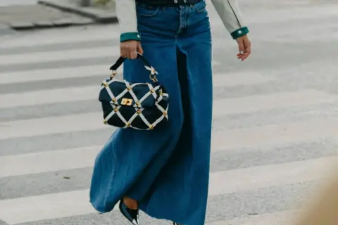 Un outfit gonna jeans lunga con borsa a tracolla trapuntata e tacchi lucidi, cammina su un attraversamento pedonale.