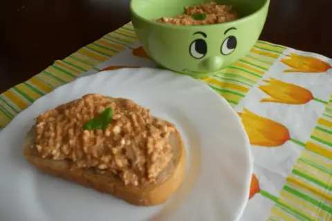Pyszna pasta z makreli i twarogu na kromce chleba, ozdobiona listkiem bazylii. Obok miseczka z pastą i wesoła buźka.