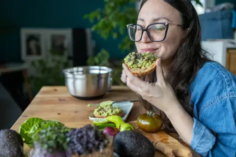 Frau genießt Avocado-Toast, ein gesunder Snack für das avocado abnehmen. Frische Zutaten liegen auf dem Tisch.