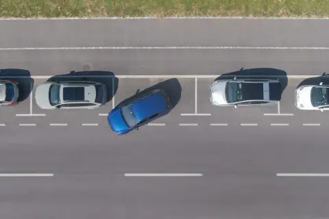 Niebieski samochód wykonuje parkowanie równoległe tyłem między dwoma innymi autami na parkingu.