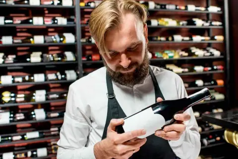 Un sommelier esamina una bottiglia, un passo fondamentale per capire se un vino è buono. Dietro di lui, scaffali pieni di bottiglie.