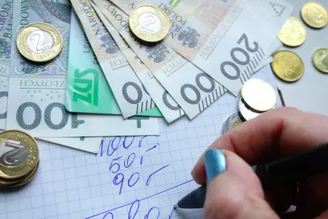 Ręka z niebieskim lakierem pisze na kratkowanym papierze, obok leżą polskie banknoty i monety. Planowanie wydatków to pierwszy krok do rewaloryzacji budżetu.