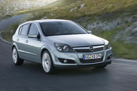 Srebrny Opel Astra 2005 na krętej drodze, otoczony zielonymi wzgórzami.