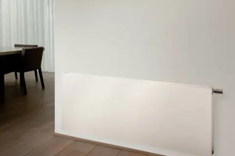Radiadores de alta temperatura blancos, modernos y discretos, instalados en una pared junto a un suelo de madera y una mesa con sillas.