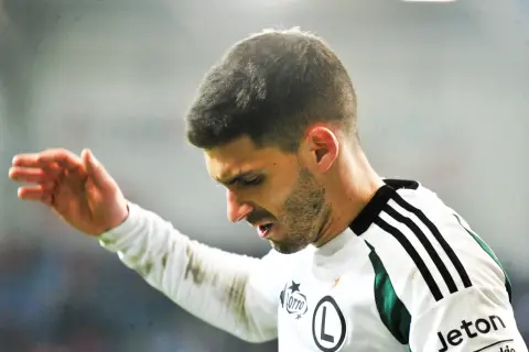 Piłkarz Legii w białej koszulce z czarnymi pasami na ramieniu, z ręką uniesioną w geście.